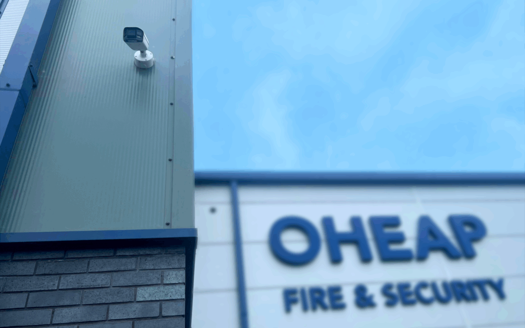 In the forefront, a CCTV camera installed to the side of a building. In the background the OHEAP sign