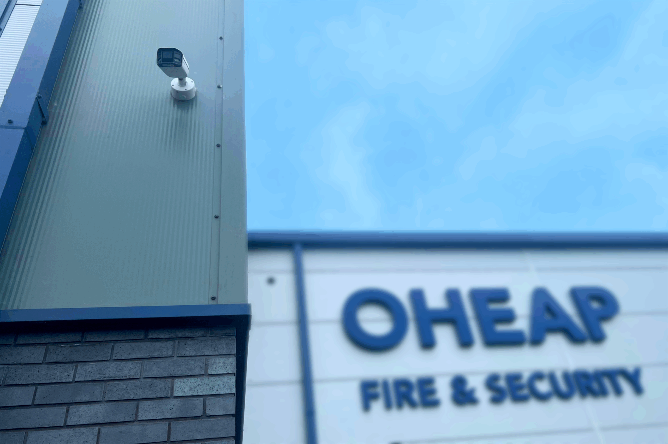 In the forefront, a CCTV camera installed to the side of a building. In the background the OHEAP sign